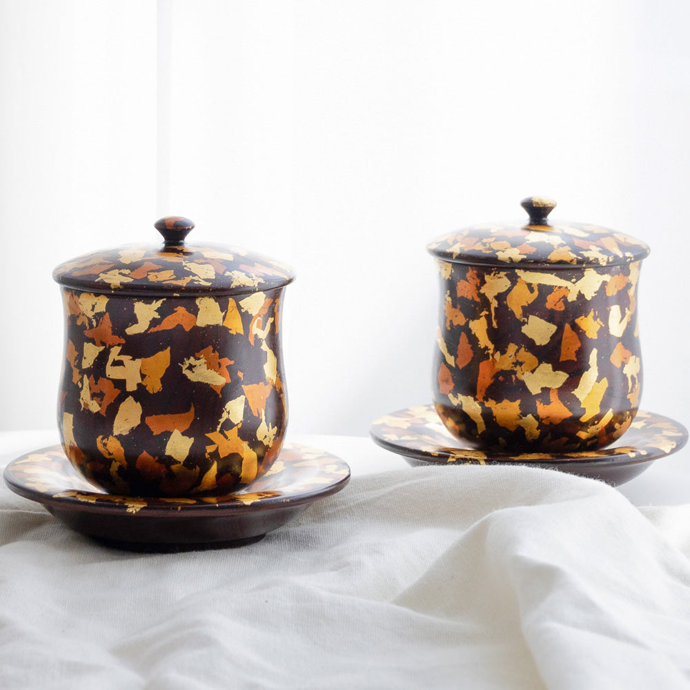 Pair of dark lacquered Korean tea cups with fitted lids on matching saucers, the surfaces scattered with irregular gold and amber flake