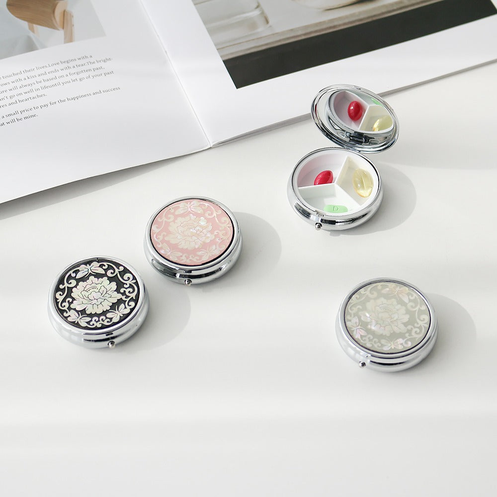Round metal Korean pill cases with lid panels of mother-of-pearl peonies on black, blush and pale jade grounds, one open showing three compartments