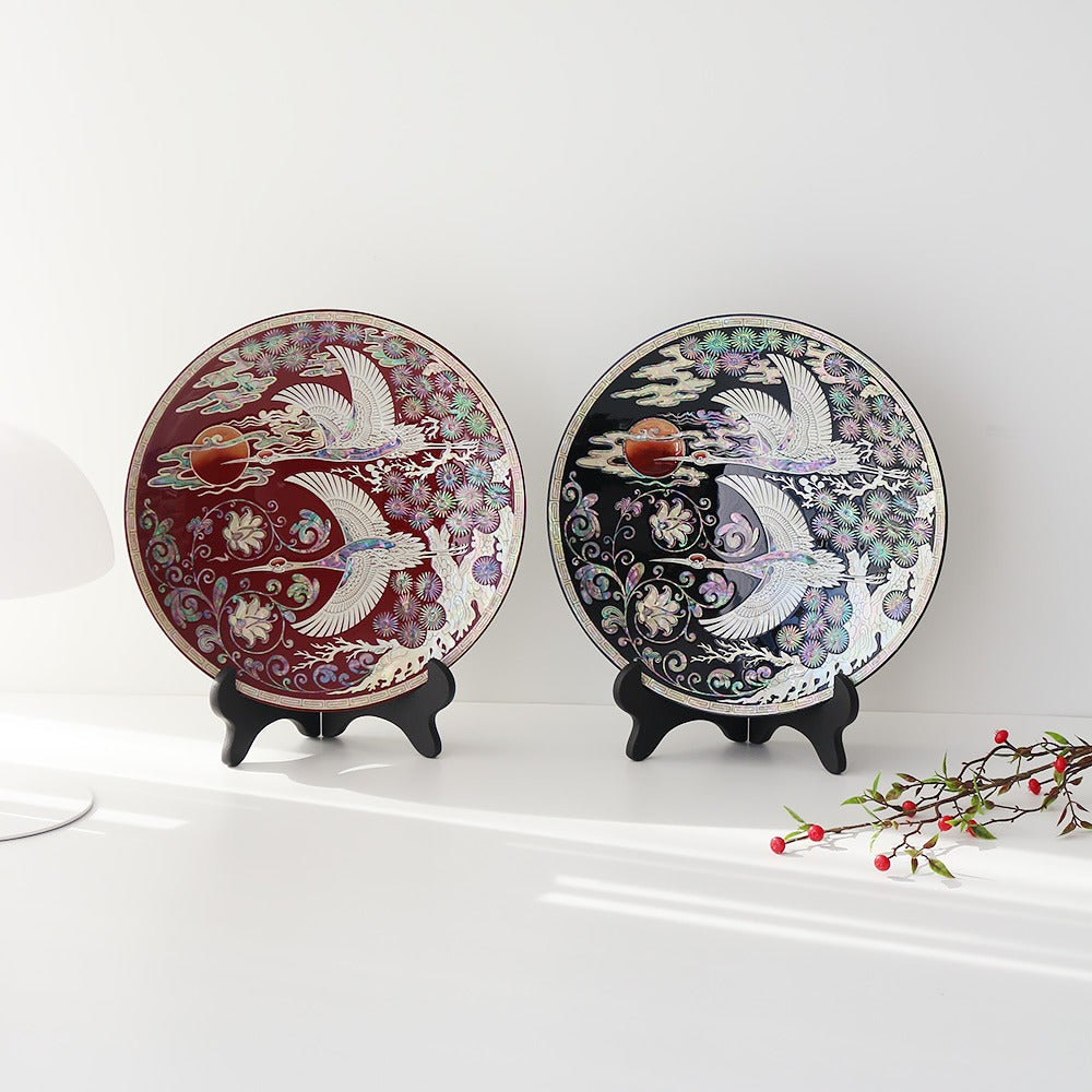 Two round Korean lacquer ornament trays in red and black on display stands, two cranes in flight beneath a round moon with pine and scrolling vines inlaid in mother-of-pearl