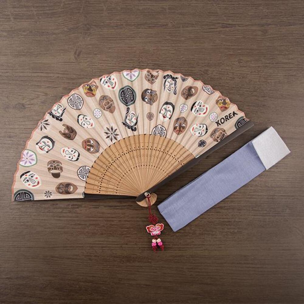 Beige silk Korean folding fan scattered with Hahoe mask faces and lattice roundels over bamboo ribs, butterfly knot tassel and a fabric sleeve beside it